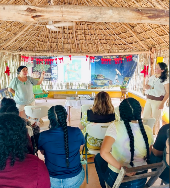 Mujeres en comunidad intercambiando saberes y experiencias educativas en un taller rural, rodeadas de niñas, niños y materiales pedagógicos.